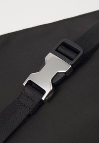 Cinghia nera in nylon con una fibbia metallica argento caratterizzata da un design a "X". Primo piano che mostra la texture del tessuto e i dettagli della fibbia.