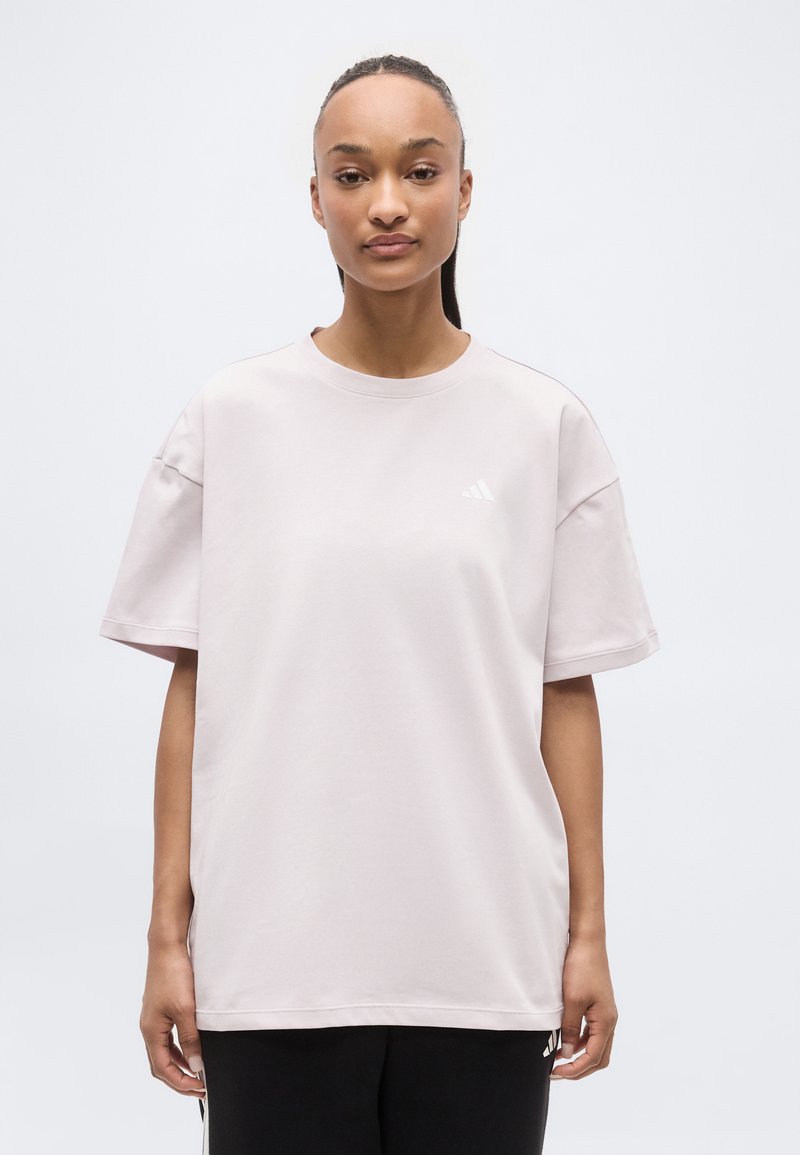 Femme aux cheveux tressés portant un T-shirt Adidas oversize beige clair et un pantalon noir, debout devant un fond blanc uni.