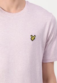 Jasnofioletowa koszula z krótkim rękawem z małym żółtym emblematem ptaka na lewej piersi, widoczna od szyi do połowy torsu.