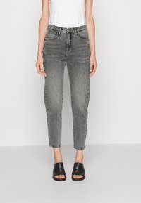 Kvinna i hög midja grå blekta jeans, vit ärmlös topp och svarta öppna sandaler med klack, stående mot en enkel bakgrund.