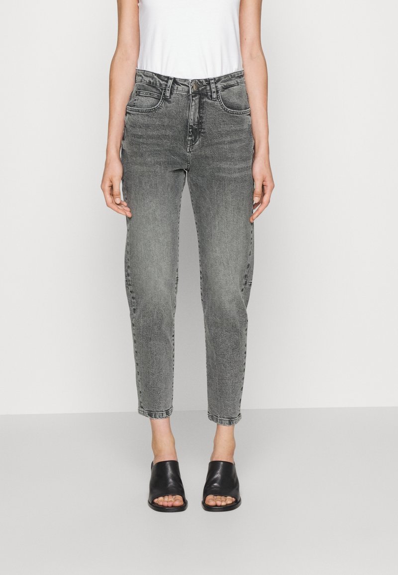 Kvinna i hög midja grå blekta jeans, vit ärmlös topp och svarta öppna sandaler med klack, stående mot en enkel bakgrund.