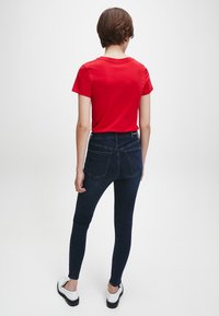 Camiseta roja de manga corta combinada con unos jeans ajustados de mezclilla oscuros de cintura alta. El modelo lleva zapatillas blancas con suelas negras. Se muestra la vista trasera.