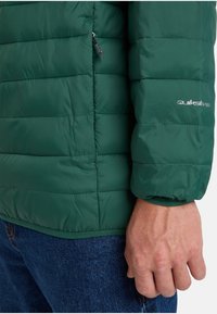 Grünes Puffermantel mit gestepptem Design, Reißverschlusstasche und Quiksilver-Logo. Glatte Textur und elastische Bündchen. Präsentiert mit blauen Jeans.