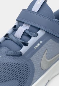 Les baskets Nike, principalement en coloris bleu, présentent une tige en maille texturée, une sangle Dynamic Fit, des lacets et un logo swoosh argenté.