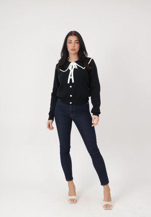 Donna in piedi, indossa un cardigan nero con colletto bianco e fiocco, jeans skinny scuri e sandali bianchi con tacco, su sfondo uniforme.