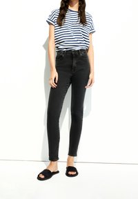 Vaqueros ajustados de talle alto negros combinados con una camiseta de rayas azules y blancas, complementados con sandalias negras de estilo deslizante. Textura de tela suave.