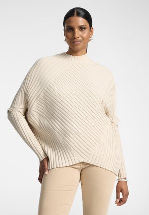 Donna con capelli scuri che indossa orecchini a cerchio e un maglione a coste crema con righe diagonali e verticali, abbinato a pantaloni beige.