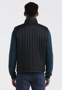 Schwarze gepolsterte Weste mit vertikalem Steppmuster, hohem Kragen und ohne sichtbare Verschlüsse, kombiniert mit einem blauen Langarmhemd.