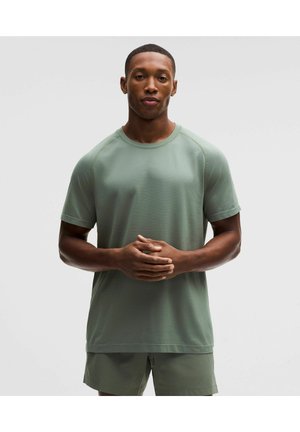 Uomo in piedi con le mani intrecciate, indossa una camicia verde chiaro a maniche corte e pantaloncini abbinati, su uno sfondo chiaro e uniforme.