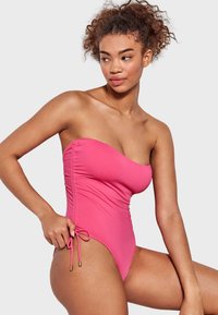 Bañador fucsia de una sola pieza con un diseño sin tirantes, detalles fruncidos en los laterales y acentos de lazo ajustables en las caderas, hecho de tela elástica.