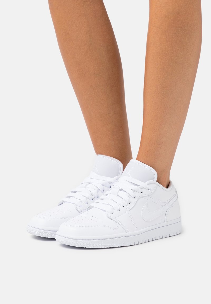 Personne portant des baskets basses blanches avec des lacets et une zone perforée au niveau des orteils, debout sur un fond clair uni.
