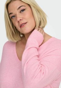 Hellrosa gestrickter Pullover mit V-Ausschnitt und gerippten Ärmeln. Die Textur wirkt weich und bietet eine lockere Passform mit dezenten Nahtdetails.