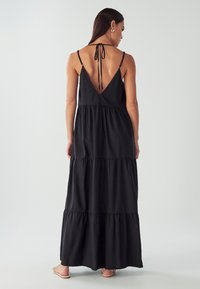 Žena v bezramínkových černých tiered maxi šatech s hlubokým V výstřihem a zavázanými ramínky, stojící bosá na jednoduchém pozadí.