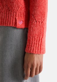 Roter gestrickter Pullover mit geripptem Muster, versehen mit einem kleinen rosa Herzen-Logo am Ärmel. Der graue Stoff der Hose ist teilweise sichtbar.