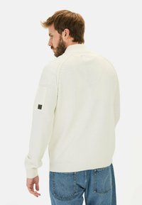 Weißer Strickpullover mit hohem Kragen, gerippten Bündchen und einem Patch an der linken Ärmels, kombiniert mit blauen Jeans.