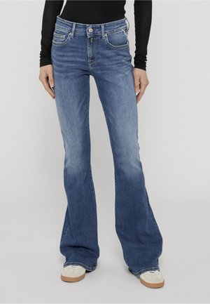 Person trägt blaue Bootcut-Jeans, weiße Sneakers und ein schwarzes Langarmshirt und steht vor einem einfarbigen hellen Hintergrund.