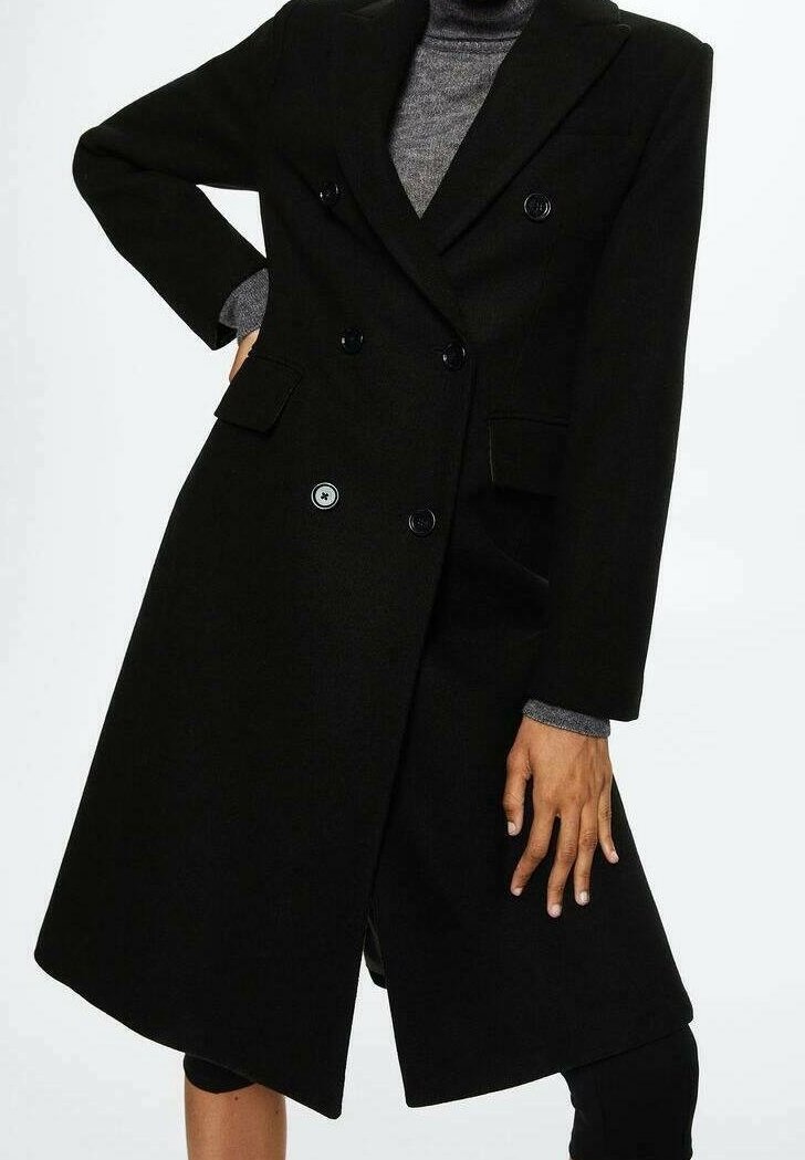 Personne portant un long manteau noir croisé par-dessus un col roulé gris, une main sur la hanche, l'autre le long du corps.