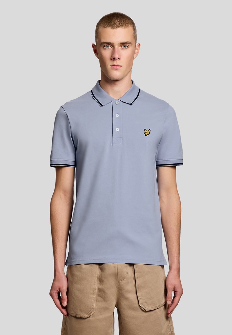 Νέος άνδρας που φοράει ανοιχτό μπλε πόλο πουκάμισο με μικρό κίτρινο λογότυπο και μπεζ παντελόνι, στέκεται μπροστά σε απλό γκρι φόντο.