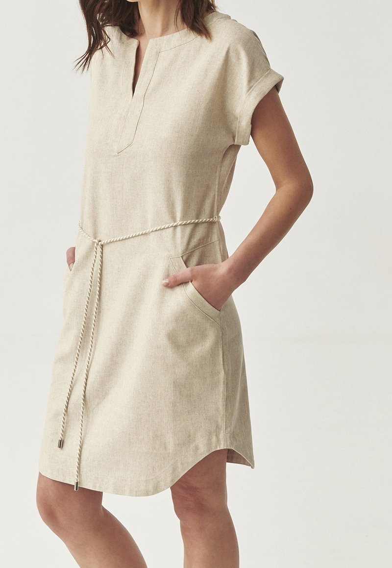 Femme portant une robe beige à manches courtes avec une ceinture en corde et des poches, debout, mains dans les poches, sur un fond clair uni.