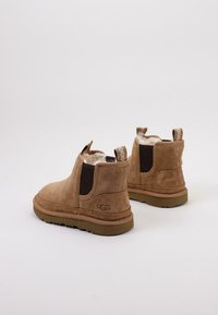 Botas de tobillo de ante marrón con un suave forro de piel de oveja, paneles laterales elásticos y una suela de goma en color neutro. Logo de UGG visible en el talón.