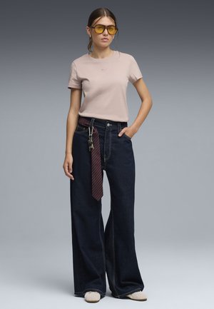 Młoda kobieta w beżowej koszulce, szerokich ciemnych dżinsach z szerokim paskiem i kluczami, w żółtych okularach przeciwsłonecznych, stoi z jedną ręką w kieszeni.