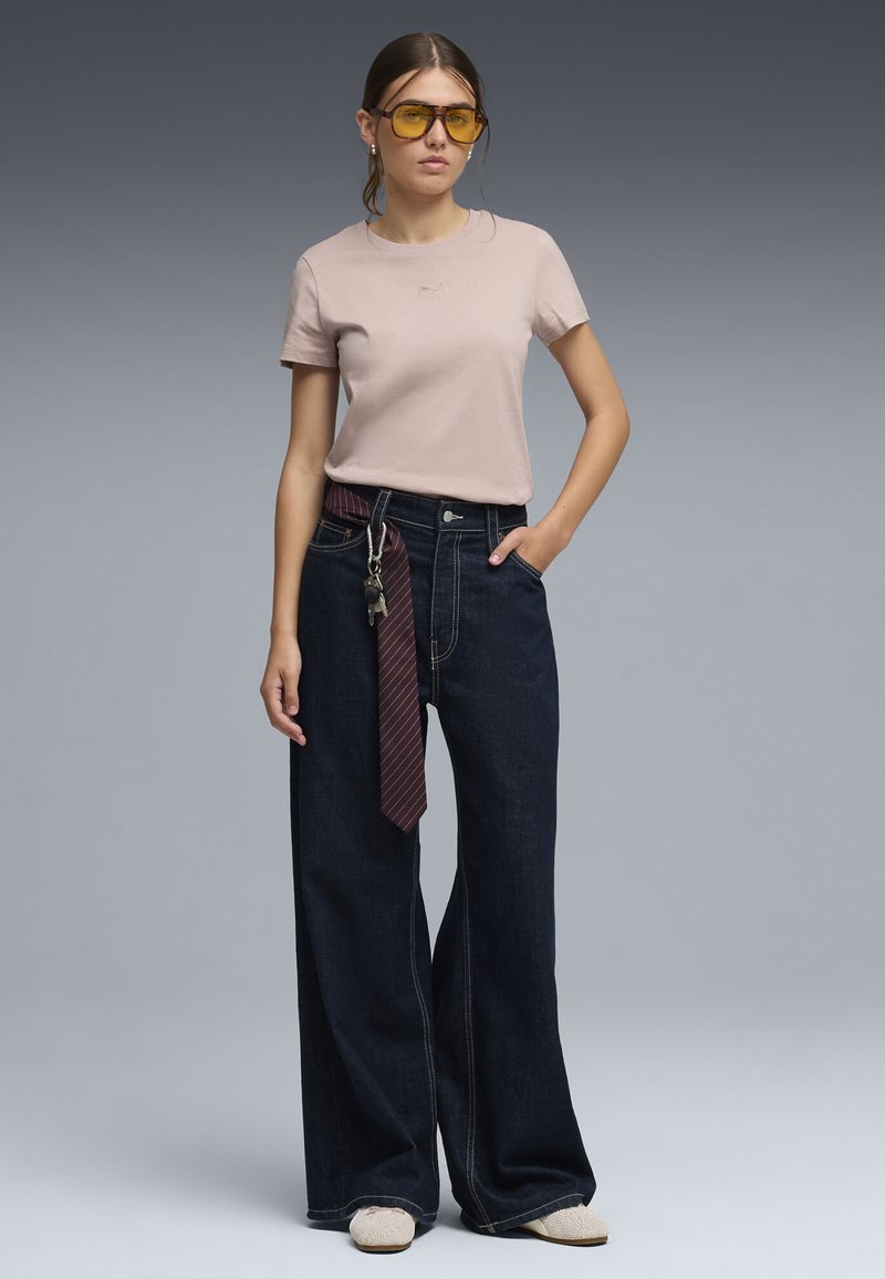 Jeune femme portant un t-shirt beige, un jean large foncé avec une ficelle et des clés suspendues, des lunettes de soleil teintées jaunes, debout avec une main dans la poche.