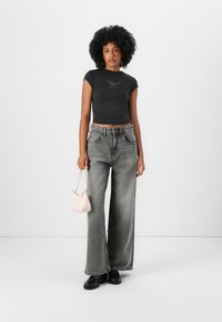 Black cropped t-shirt with wing design, paired with wide-leg gray jeans and a small pink handbag. Model wears black shoes.