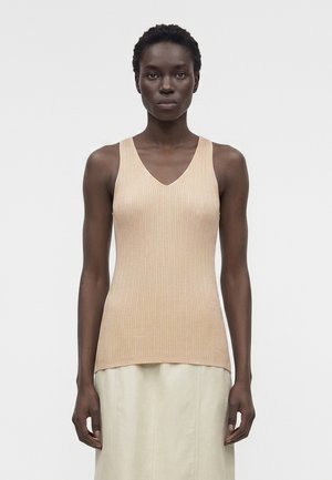 Γυναίκα με κοντά σγουρά μαλλιά που φοράει μπεζ τοπ χωρίς μανίκια με ριμπ ύφανση και φούστα ανοιχτού χρώματος, στέκεται μπροστά σε απλό ανοιχτό φόντο.