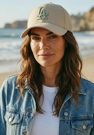 Frau mit langen, welligen Haaren, die eine beige LA-Kappe, ein weißes Hemd und eine Jeansjacke trägt und an einem Strand mit Ozean und Klippen im Hintergrund steht.