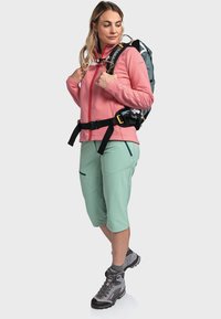 Rosa Fleecejacke mit einem vollständigen Reißverschluss, grüne Shorts mit Taschen und graue Wanderschuhe. Der Rucksack hat schwarze Riemen und Beschläge.
