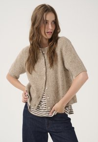Lysbrun kortærmet cardigan med en blød tekstur, udstyret med runde perleknapper. Bærer den over en grå og hvid stribet top samt mørke jeans.