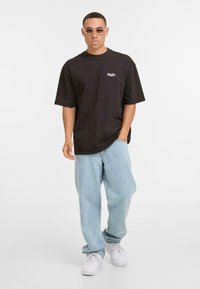 Sort bomulds oversized t-shirt med korte ærmer, parret med lyseblå denimjeans og hvide sneakers; modellen bærer klare solbriller.