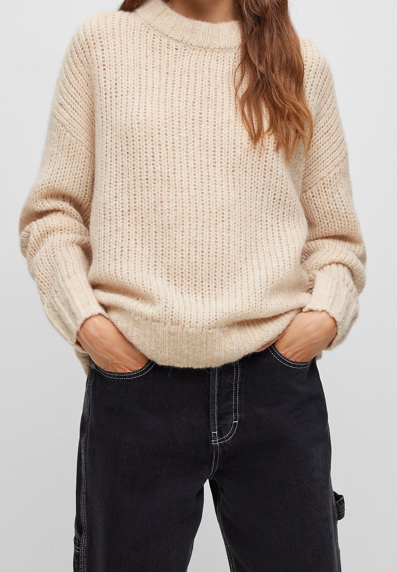 Femme portant un pull en grosse maille crème avec les mains dans les poches d'un jean noir taille haute avec des détails de couture blanche.