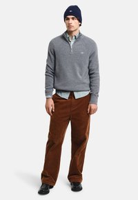 Grauer Strickpullover mit Reißverschlusskragen über einem hellblauen und weißen gestreiften Hemd. Braune Cordhosen und eine dunkelblaue Strickmütze.