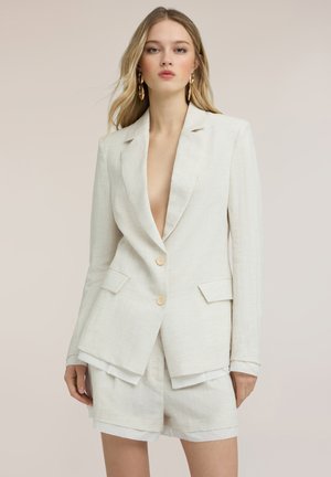 Femme blonde portant un blazer crème oversize avec deux boutons et un short assorti, debout devant un fond beige.