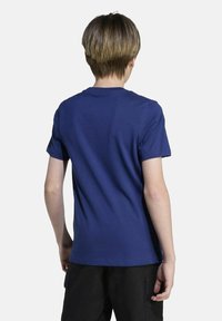 Persoon met kort bruin haar, gekleed in een effen blauwe T-shirt met korte mouwen en zwarte broek, staande met de rug naar de camera op een witte achtergrond.