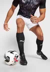 Maglia a strisce nere e grigie, pantaloni corti bianchi, calze nere e scarpini da calcio neri; un pallone da calcio a fantasia sul terreno.