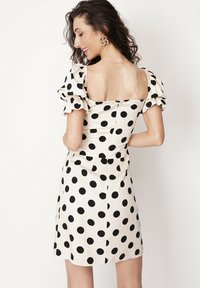 Kremowa sukienka w czarne kropki, z wyraźnym dekoltem, krótkimi bufiastymi rękawami, dopasowaną talią i lekko rozkloszowanym dołem. Zamek z tyłu.
