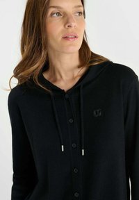Zwarte hoodie cardigan met een frontale knoopsluiting, voorzien van een klein logo, gemaakt van een gestructureerde stof. Zacht, casual ontwerp met trekkoorden.
