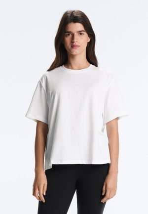 Mladá žena s dlhými hnedými vlasmi, oblečená v voľnom bielom tričku a čiernych nohaviciach, stojaca pred jednoduchým svetlým pozadím.