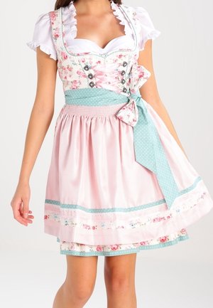 Femme portant une robe dirndl fleurie rose et blanche avec une ceinture bleue à pois nouée en nœud à la taille.