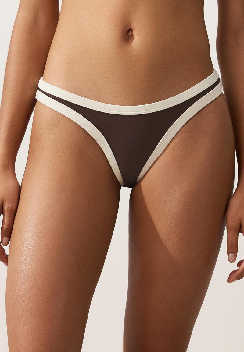 Femme portant un bas de bikini marron avec bord blanc, montrée de la taille jusqu'à mi-cuisse sur un fond uni.