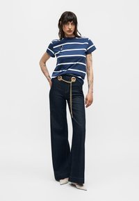 Γυναίκα με μπλε ναυτικό και λευκό ριγέ μπλουζάκι, σκούρο τζιν με φαρδιά μπατζάκια και χρυσή αλυσίδα στη μέση και λευκά μυτερά παπούτσια, στέκεται μπροστά σε μονόχρωμο φόντο.