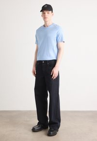 Light blue cotton t-shirt, dark blue denim jeans, black cap with white text, black chunky shoes, standing on a neutral-colored floor.
