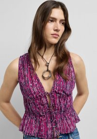 Jonge vrouw met lang bruin haar, een paarse top zonder mouwen met een patroon en een zwarte koordketting met een zilveren ronde hanger.