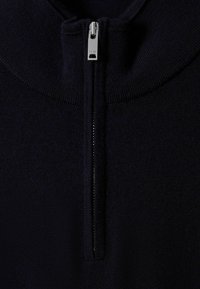 Pull en mailles bleu marine avec col haut et zip central. Il présente une texture lisse et des accents côtelés près du col.