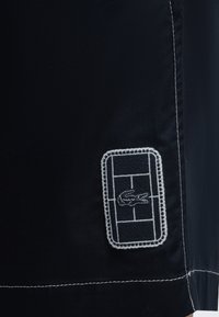 Tissu noir avec un patch rectangulaire blanc cousu montrant un court de tennis et un logo de crocodile au centre.