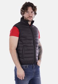 Gilet imbottito nero con colletto alto, chiusura a zip e cuciture orizzontali. Indossato sopra una camicia rossa, con finitura testurizzata e tasche.