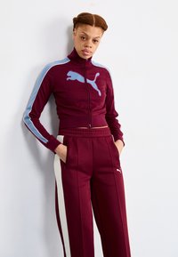 Bordeaux trainingspak met lichtblauwe accenten, voorzien van een hoge kraag en geborduurd logo op de borst. De broek heeft ritszakken en zijstrepen.
