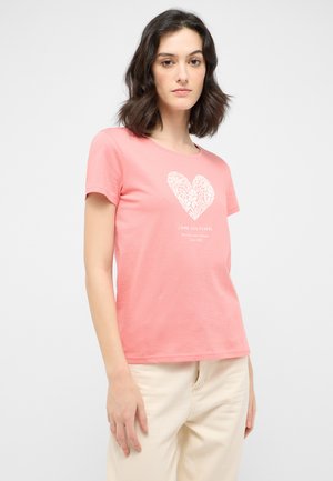 T-shirt en coton rose à manches courtes, avec un dessin de cœur blanc et le texte "J'aime les fleurs" en dessous. Porté avec un pantalon clair.